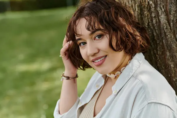 A cheerful young woman in a white shirt relaxes under a tree in a vibrant summer park. — Stock Photo