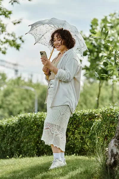 A cheerful young woman in a white shirt smiles while using her phone under a delicate umbrella in summer. — Stock Photo