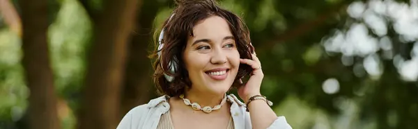 A cheerful young woman in a white shirt relaxes in a lush green park, fully immersed in her music. — Stock Photo