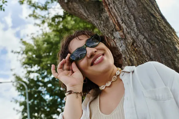 A cheerful young woman wearing a white shirt playfully adjusts her sunglasses under a vibrant summer sky. — Stock Photo