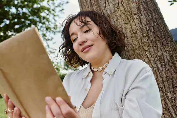 A young woman in a white shirt enjoys a peaceful moment reading under a tree in a vibrant park. — Stock Photo