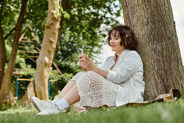 A young woman in a white shirt enjoys her summer day, engaging with her phone under a leafy tree. — Stock Photo