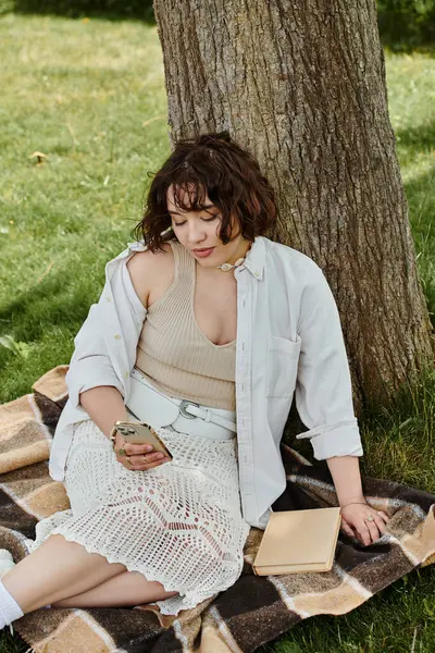 A pretty young woman relaxes in a green park during summer, engrossed in her phone while seated on a cozy blanket. — Stock Photo