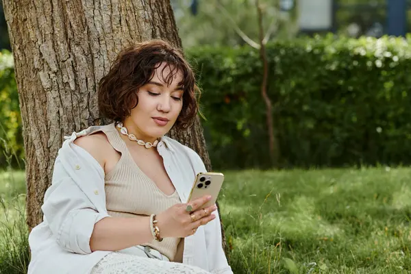 A young woman relaxes under a tree, soaking up the summer sun while scrolling through her phone. — Stock Photo
