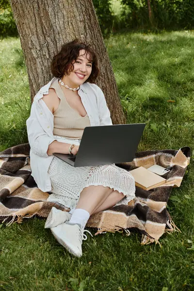 A cheerful young woman types on her laptop, relaxed on a blanket in a sunny park filled with greenery. — Stock Photo