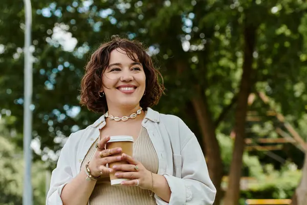 A cheerful young woman in a white shirt relaxes in a vibrant park, savoring her coffee under the summer sun. — Stock Photo