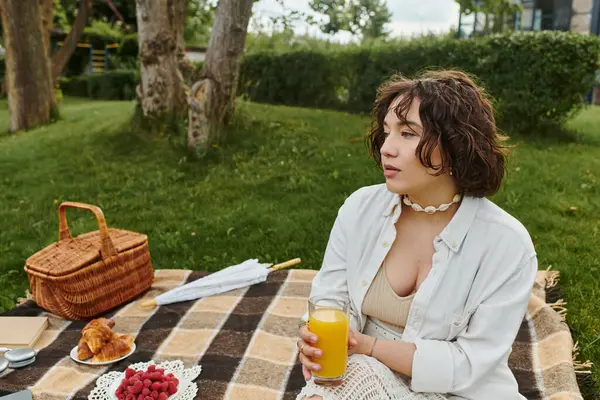 A young woman enjoys a peaceful moment, sipping juice while resting on a picnic blanket surrounded by nature. — Stock Photo