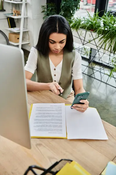 Concentrated woman reviews documents while checking her phone in a vibrant workspace. — Stock Photo