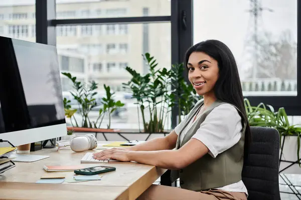 A poised woman engages with her computer in a vibrant workspace. — Stock Photo