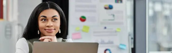 A poised woman reflecting while using her laptop in a vibrant workspace. — Stock Photo