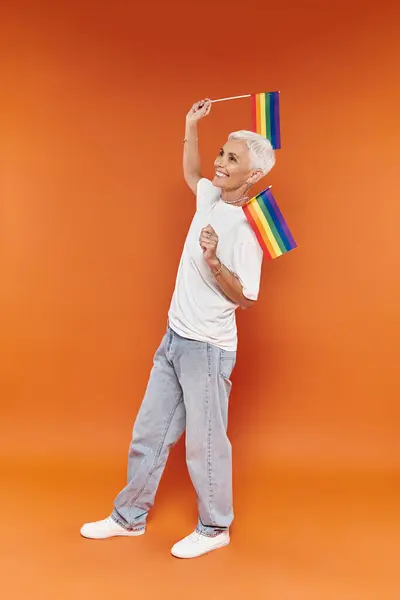 A mature woman with short hair joyfully waves rainbow flags in celebration. — Stock Photo
