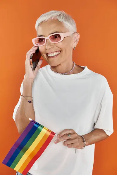 A mature woman joyfully connects on the phone, holding a pride flag. — Stock Photo