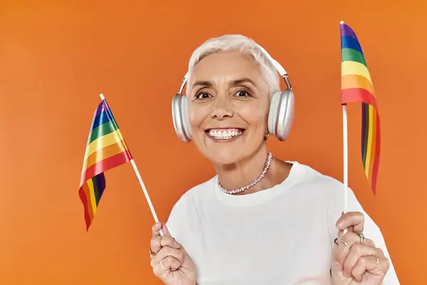 Mature woman with short hair joyfully waves rainbow flags while listening to music. — Stock Photo