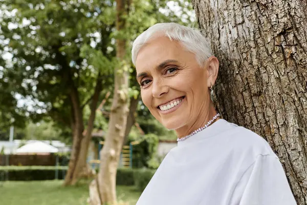 A mature woman beams with joy while leaning against a tree. — Stock Photo