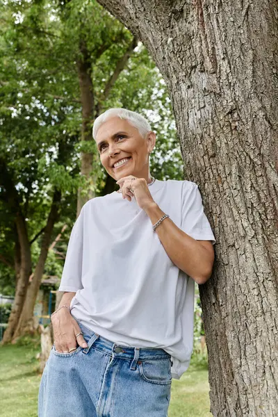 Mature woman smiles while leaning against a tree, embracing a sunny day outdoors. — Stock Photo