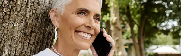 A mature woman with short hair smiles while on a phone call outdoors. — Stock Photo