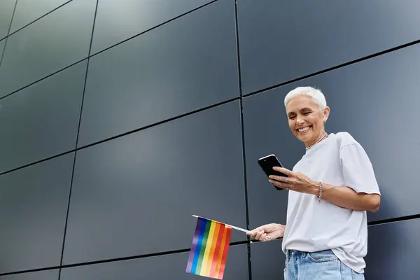 Donna sorridente tiene una bandiera arcobaleno mentre scrive in una città moderna. — Foto stock