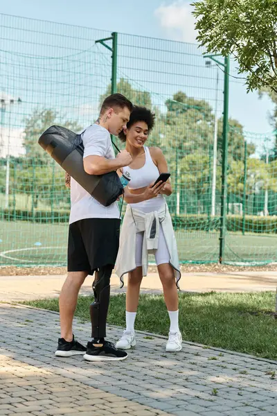 A young woman and a tattooed man with a prosthetic leg share workout tips, looking at smartphone in a sunny park. — Stock Photo