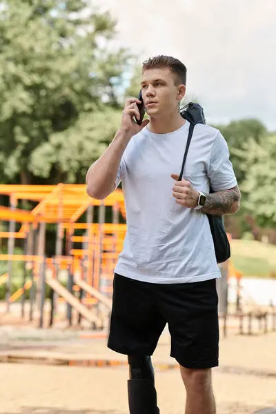 Un hombre tatuado con una pierna protésica, con una camiseta blanca y pantalones cortos negros, camina mientras habla por teléfono. — Stock Photo