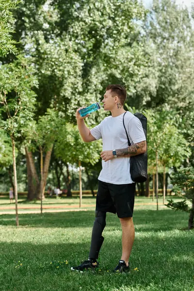 A young man with tattoos sips water in a lush green park, embracing inclusion and outdoor adventure. — Stock Photo