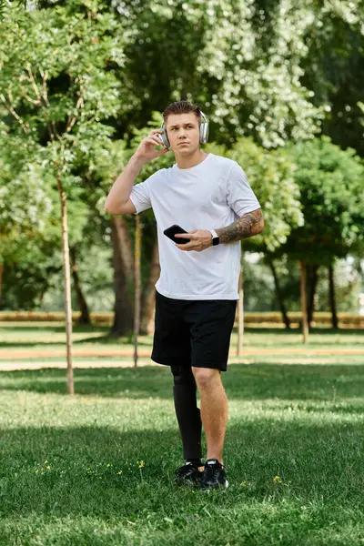 Joven tatuado con pierna artificial escuchando música y usando smartphone en un parque soleado - foto de stock