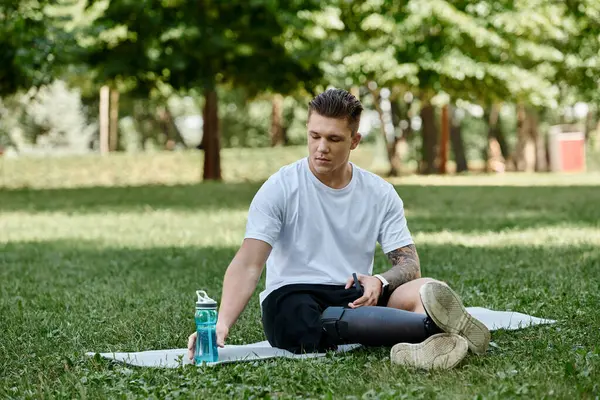 Tattooed young man with prosthetic leg sitting on fitness mat in park, reaching for water — Stock Photo