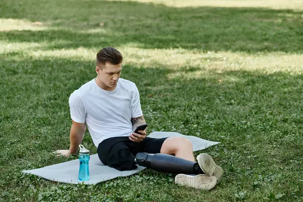 Tattooed young man with prosthetic leg sitting on fitness mat and using smartphone in park — Stock Photo