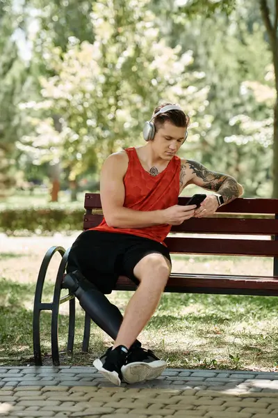 A young man with tattoos sits on a bench, focused on his phone, enjoying the music through headphones. — Stock Photo