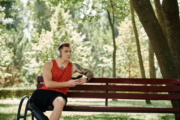 A young man with tattoos sits on a bench outdoors, focused on his phone, enjoying the music through headphones. — Stock Photo