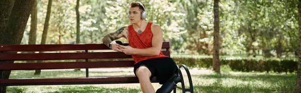 Un joven con tatuajes se sienta en un banco al aire libre, disfrutando de la música a través de auriculares. - foto de stock
