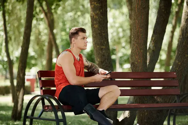 Un jeune homme tatoué est assis sur un banc en bois dans un parc verdoyant à l'extérieur — Photo de stock