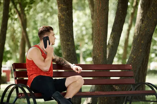 A young man with tattoos sits on a bench and having phone call in green park outdoors — Stock Photo