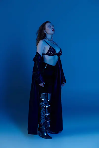 A confident woman with red hair poses in trendy attire against a deep blue backdrop. — Stock Photo