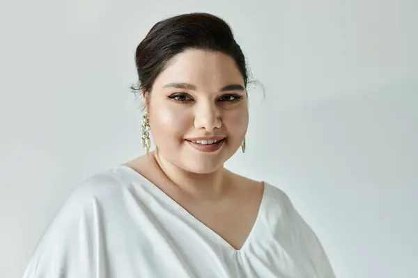A stunning plus size bride radiates joy, adorned in a white gown and sparkling earrings on a serene grey background. — Stock Photo