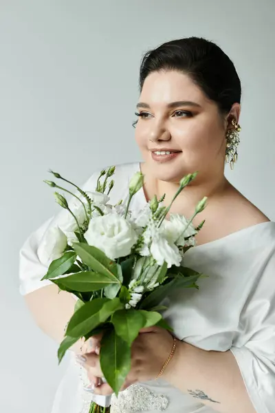 A stunning plus size bride in a white dress embraces a bouquet of white flowers against a subtle gray backdrop. — Stock Photo