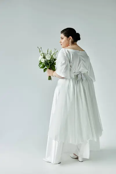 A beautiful plus size bride stands gracefully in a flowing white gown, holding a bouquet against a grey backdrop. — Stock Photo