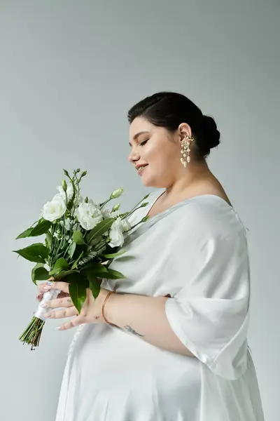 Красивая невеста plus size восхищается своим цветочным букетом в потрясающем белом платье. — стоковое фото