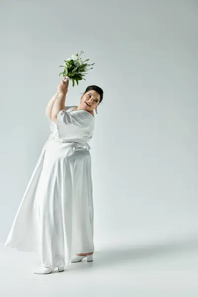 Una sposa radiosa in uno splendido abito bianco travi mentre tiene un bouquet lussureggiante su uno sfondo grigio morbido. — Foto stock