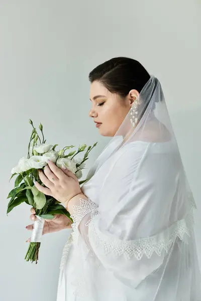 A stunning plus-size bride lovingly admires her bouquet while adorned in a beautiful white gown and veil. — Stock Photo