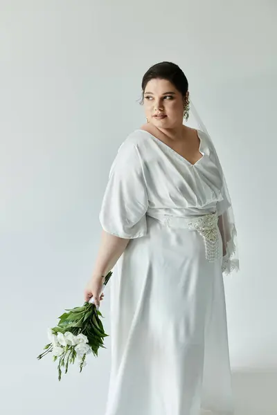 A plus-size bride in a flowing white gown holds vibrant flowers, radiating elegance against a soft grey backdrop. — Stock Photo