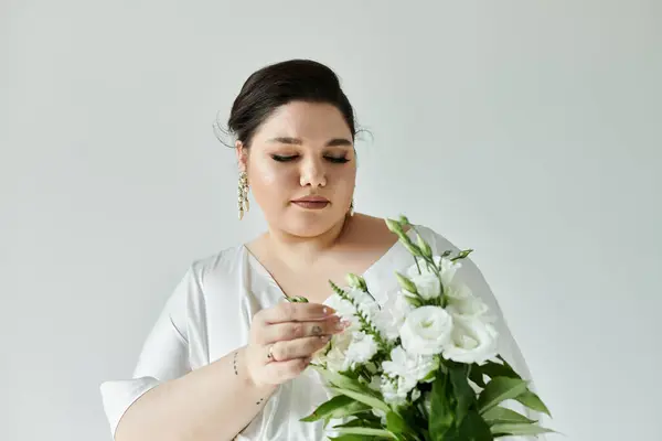 Uma jovem noiva deslumbrante em um vestido fluindo ajusta suavemente seu buquê contra um fundo cinza macio. — Fotografia de Stock