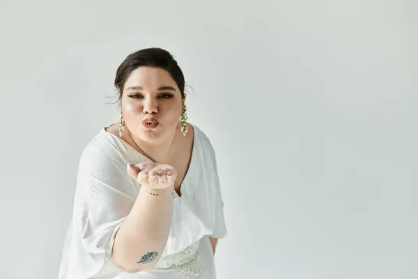 A joyful plus-size bride in a white gown blows a kiss, radiating beauty and confidence against a muted grey backdrop. — Stock Photo