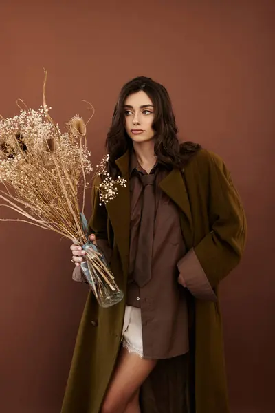 Uma jovem mulher posa elegantemente em um olhar de outono elegante, segurando flores secas contra um pano de fundo quente. — Fotografia de Stock