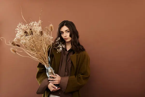 Une jeune femme élégante pose élégamment en tenue d'automne, tenant un bouquet de fleurs séchées sur un fond chaud. — Photo de stock