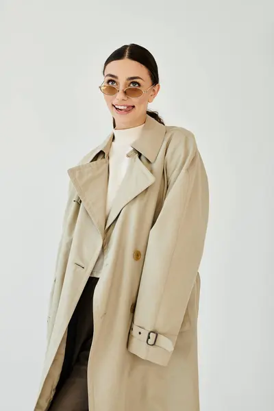 A young woman showcases her autumn style, wearing a trendy trench coat and smiling confidently in a studio. — Stock Photo