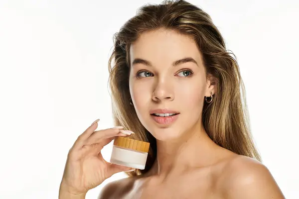 A young woman highlights her radiant skin, holding a skincare jar, radiating confidence and beauty. — Stock Photo