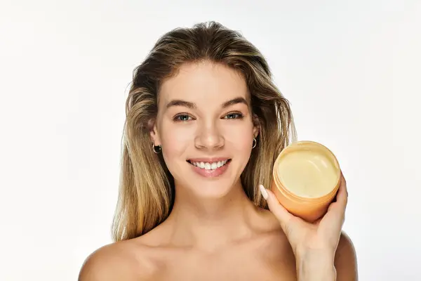 A young woman beams with joy while showcasing a jar of skin cream against a luminous background. — Stock Photo