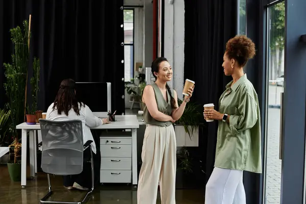 Two professional women chat and enjoy coffee while a colleague works at a desk nearby. — Stock Photo