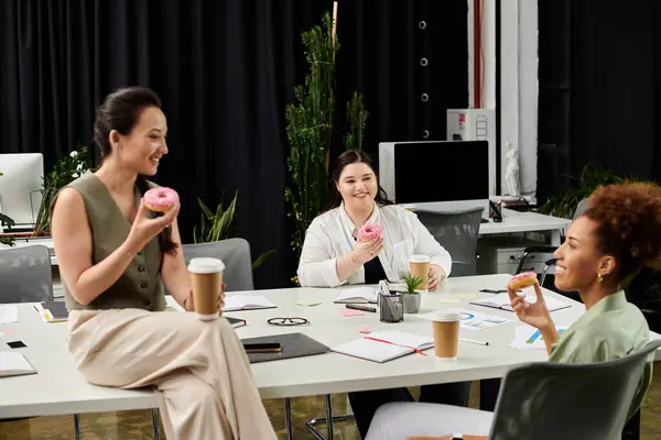 Three professional women share a lighthearted moment over coffee and donuts in an office setting. — Stock Photo