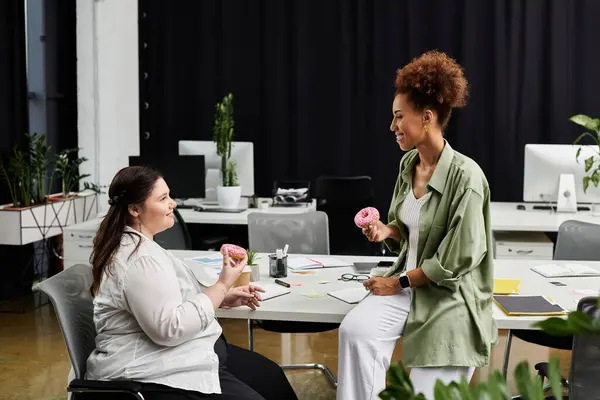 Two stylish pros chat animatedly while enjoying donuts in a trendy office. — Stock Photo